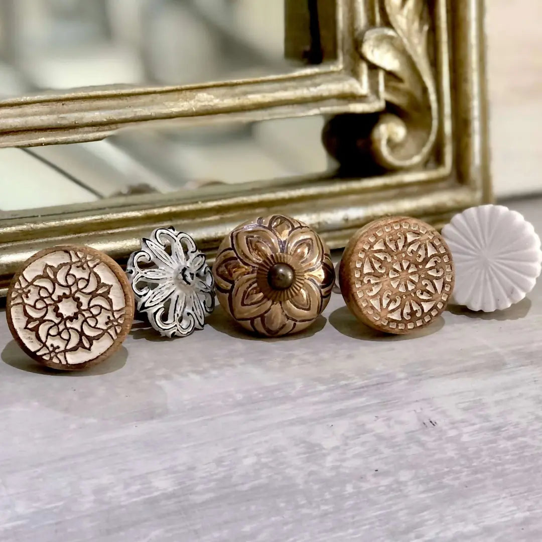 A styled display showing five decorative cabinet knobs, including the Zoe white and wood carved knob, arranged on a light wooden surface with a gold‑framed mirror behind.