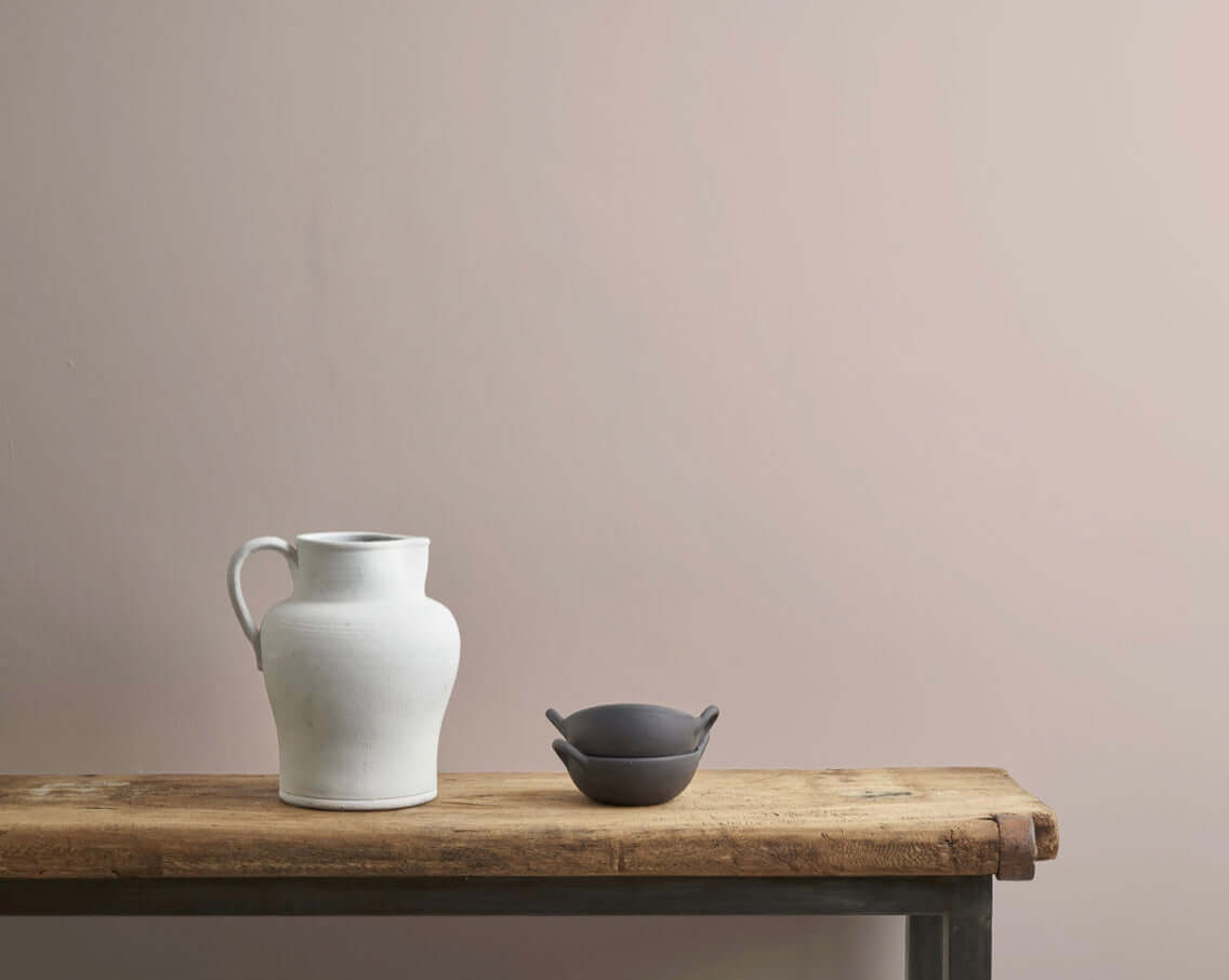 Minimalist decor featuring a white ceramic jug and a dark bowl on a wooden table against a soft pink wall.