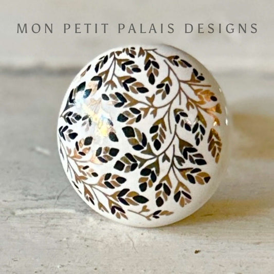 A white ceramic knob with black and gold dainty leaf motifs.