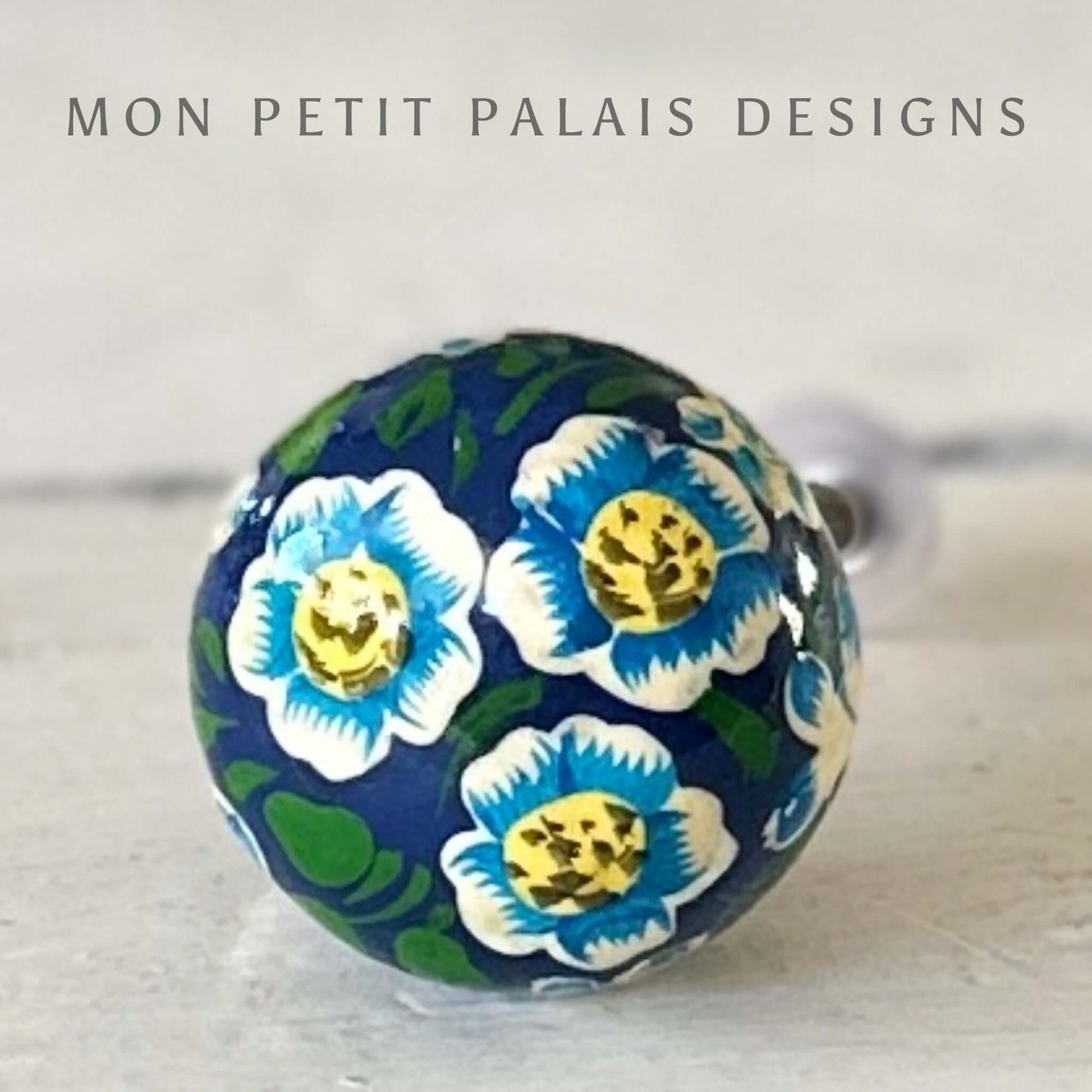A decorative spherical knob with a floral design in blue, green, and white colors, branded by MON PETIT PALAIS DESIGNS.