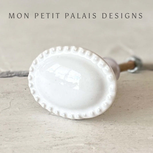 A white vintage ceramic knob with an oval shape and raised detail edge.