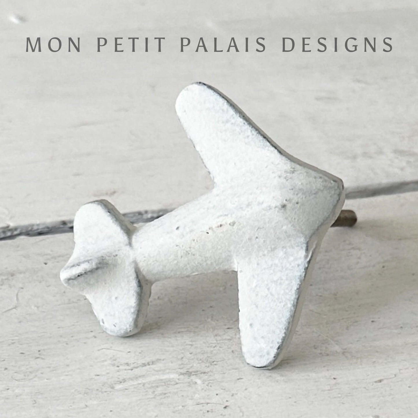 A white vintage metal knob designed to resemble an aeroplane, with a textured surface, mounted on a flat surface.
