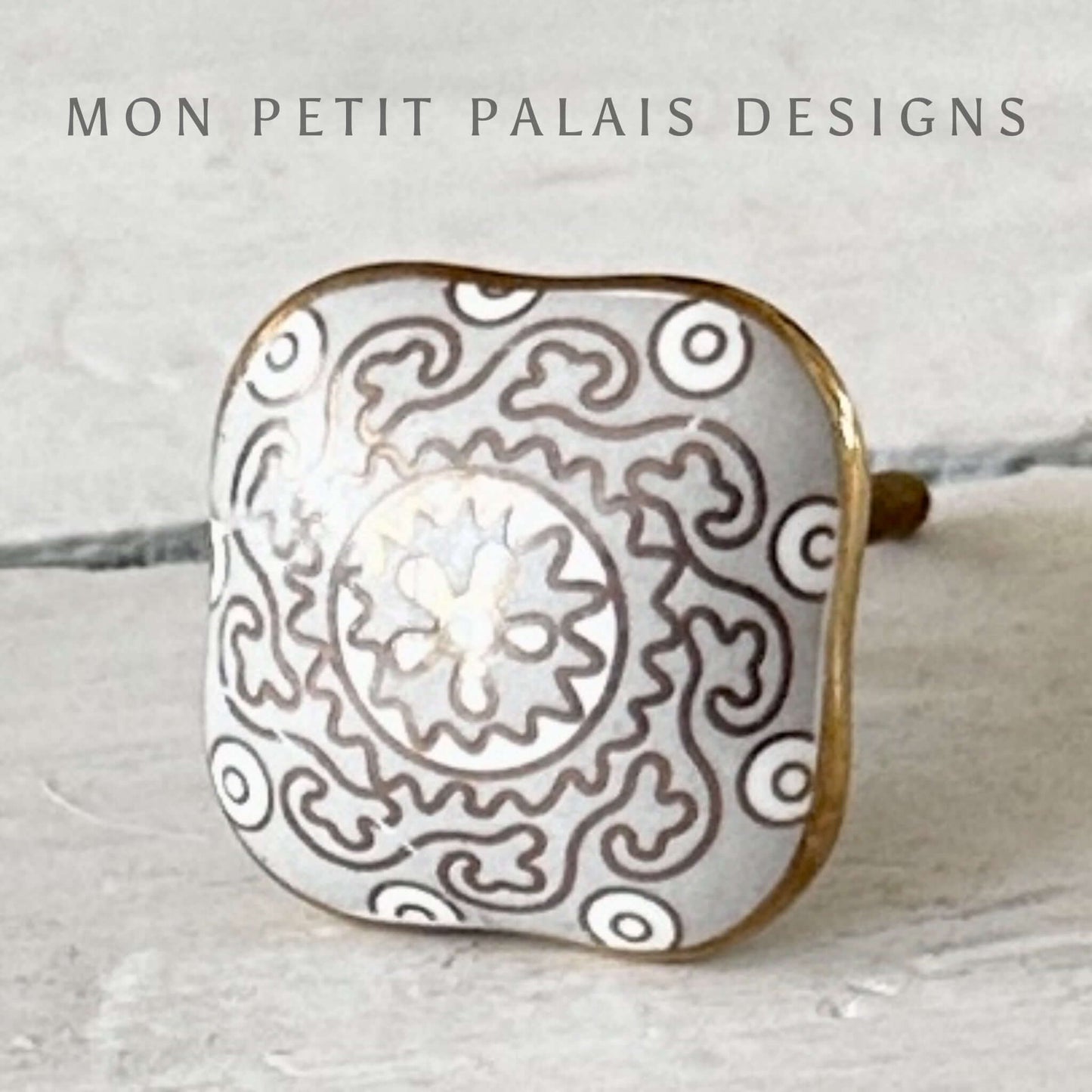A decorative ceramic knob with a geometric pattern in a light grey and gold color scheme, branded by MON PETIT PALAIS DESIGNS.