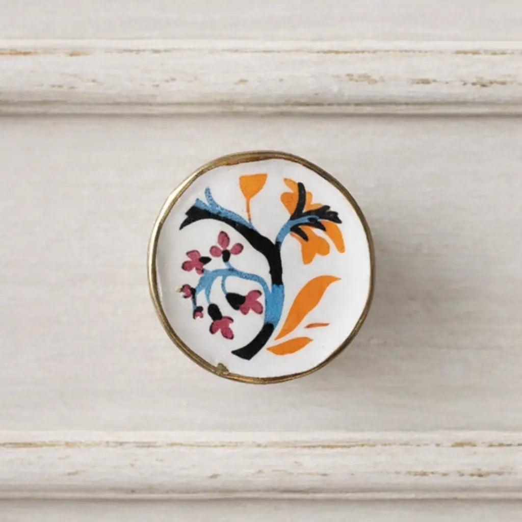 A close-up of the Soleil hand-painted ceramic cabinet knob mounted on a distressed off-white drawer front, featuring a white base with a brass rim and a colourful floral tree motif in blue, orange, black, and pink.