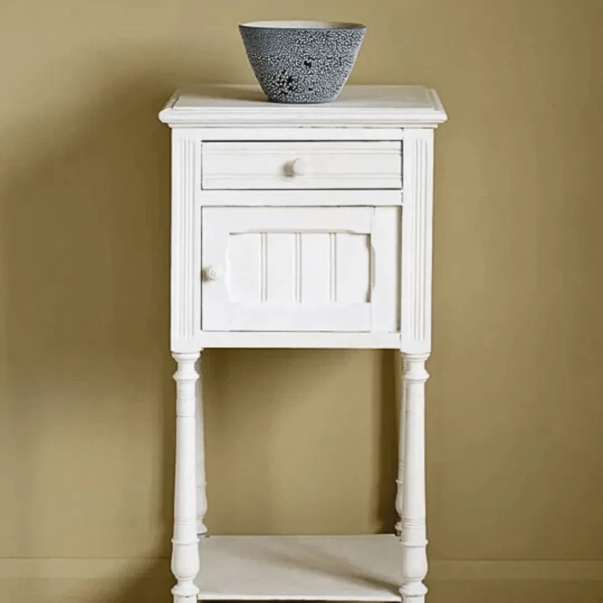 White wooden side table with a single drawer and a decorative bowl on top against a beige wall.