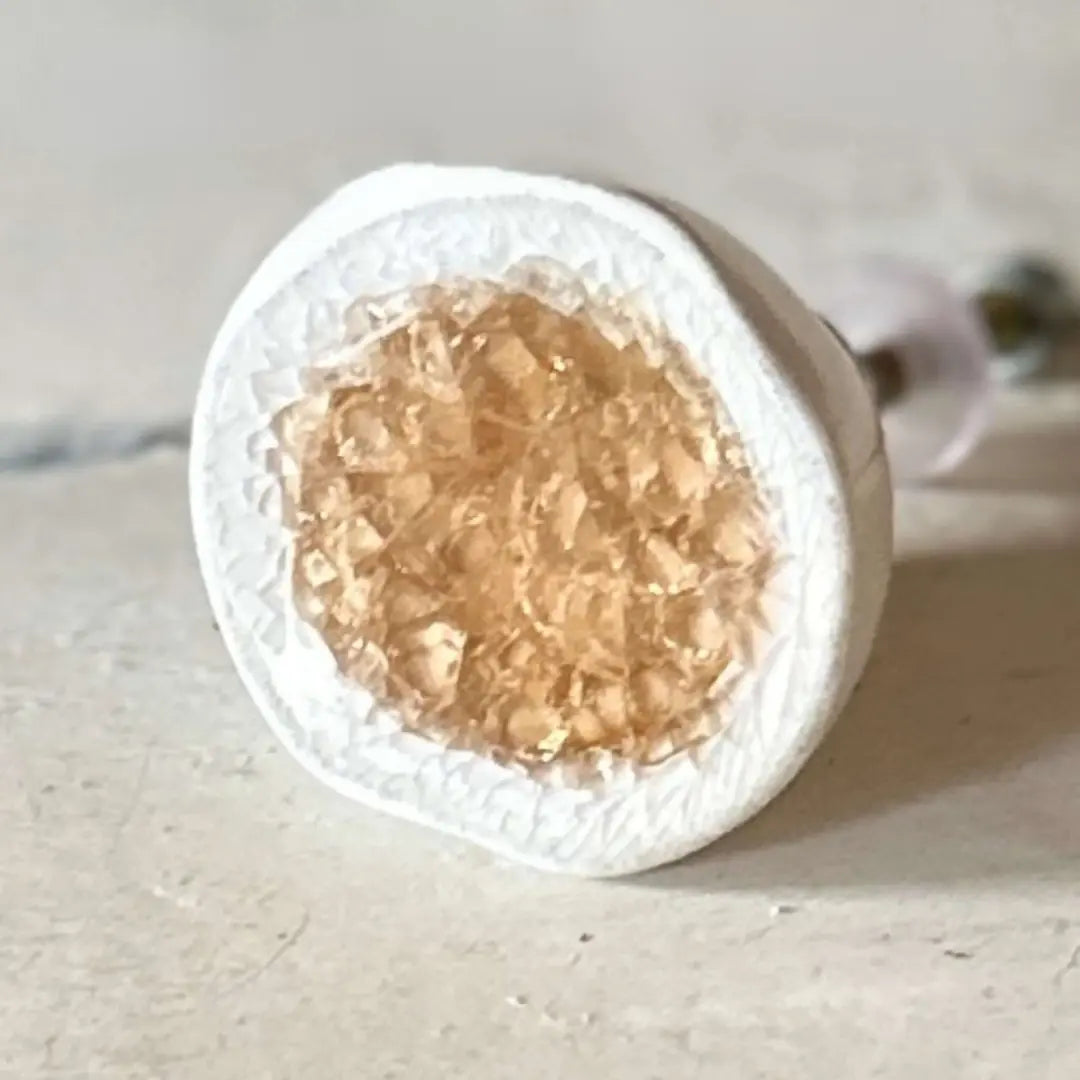 A close-up of a ceramic toffee and white ceramic knob with a translucent amber-gold crystalline center, photographed on a light neutral surface.