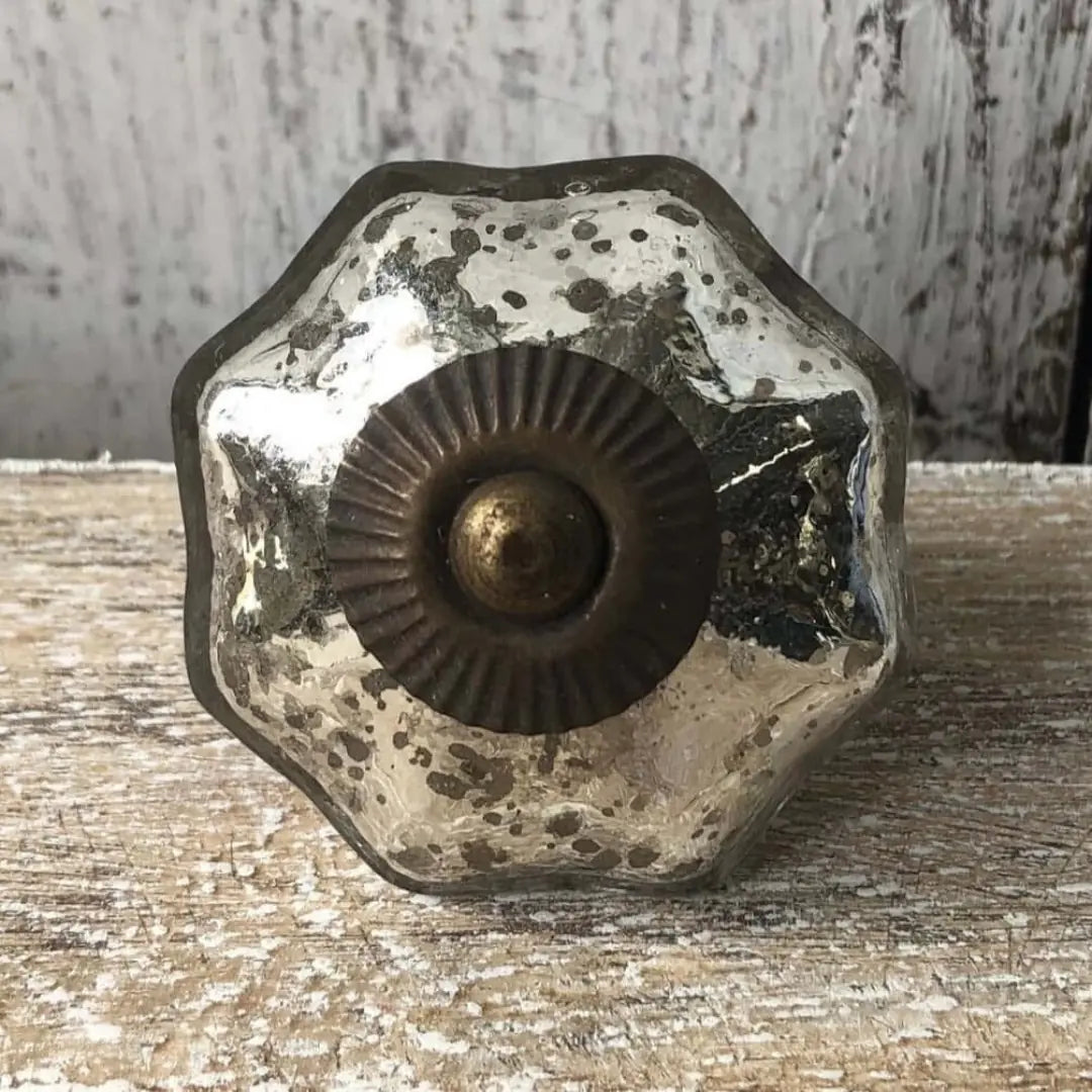 A close-up of the Vivienne mercury glass knob resting on a rustic wooden surface, showing its faceted flower shape, speckled finish, and decorative brass center.