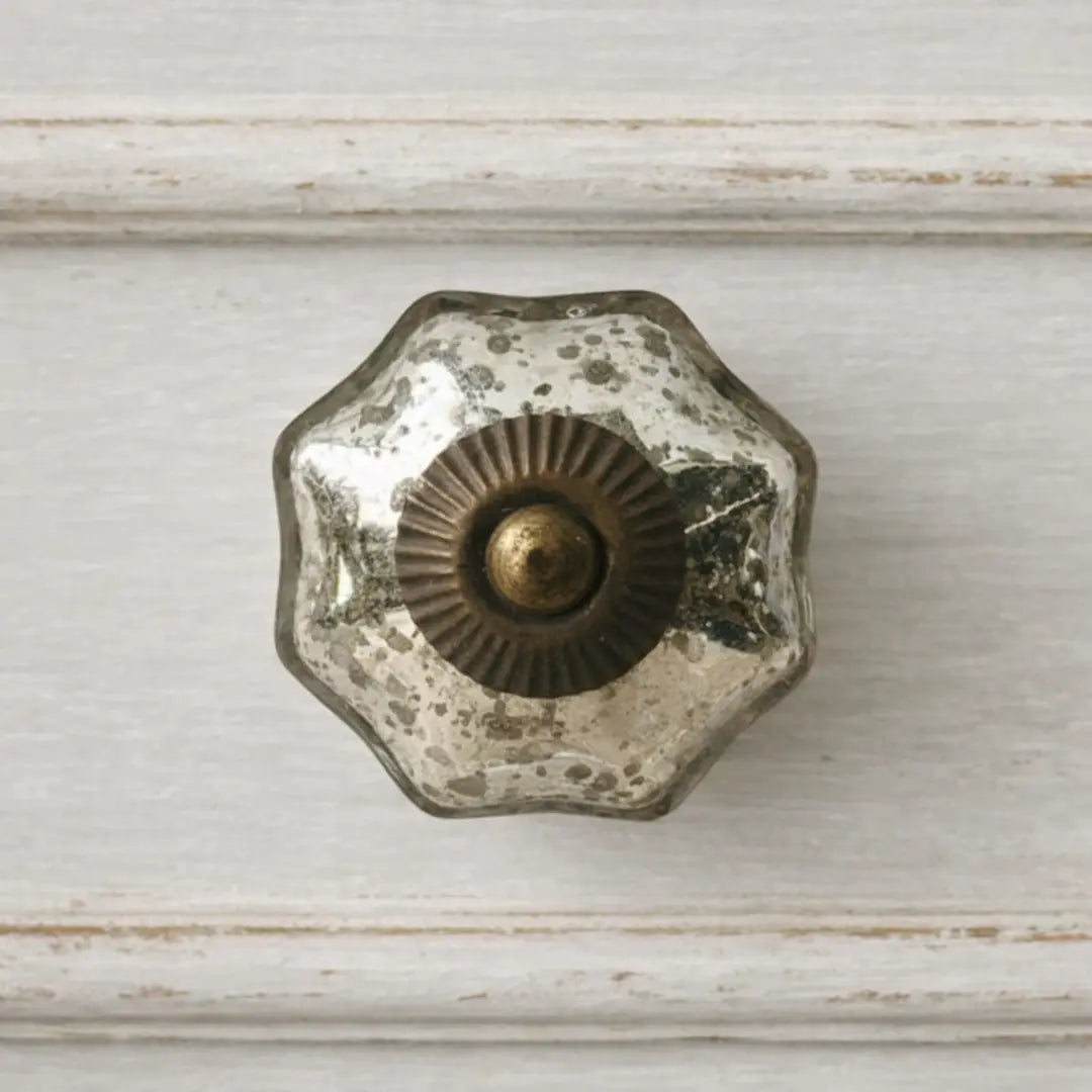 A front‑on view of the Vivienne mercury glass cabinet knob mounted on a distressed off‑white drawer, featuring a scalloped flower shape, speckled silver‑gold finish, and antique brass sunburst center.
