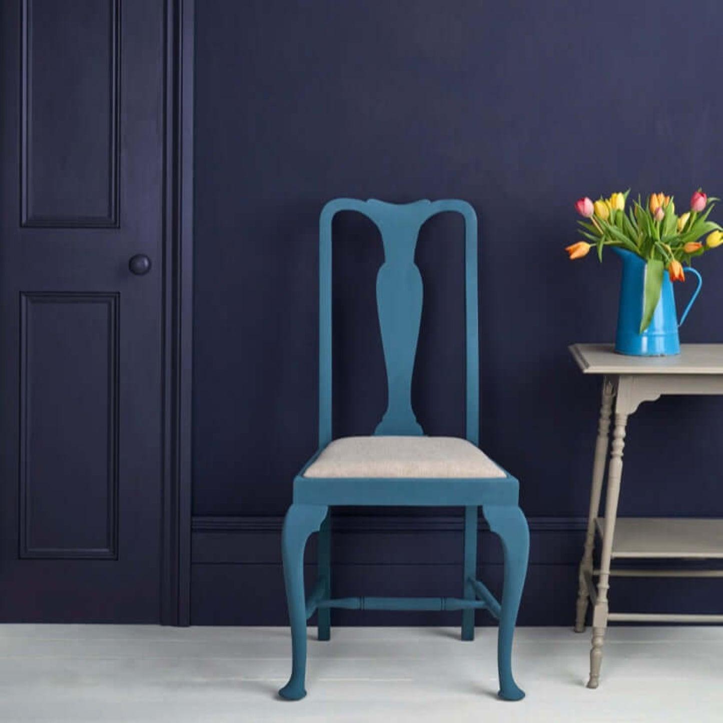 A stylish chair painted in Oxford Navy - Annie Sloan Satin Paint against a dark wall, with a vase of tulips.
