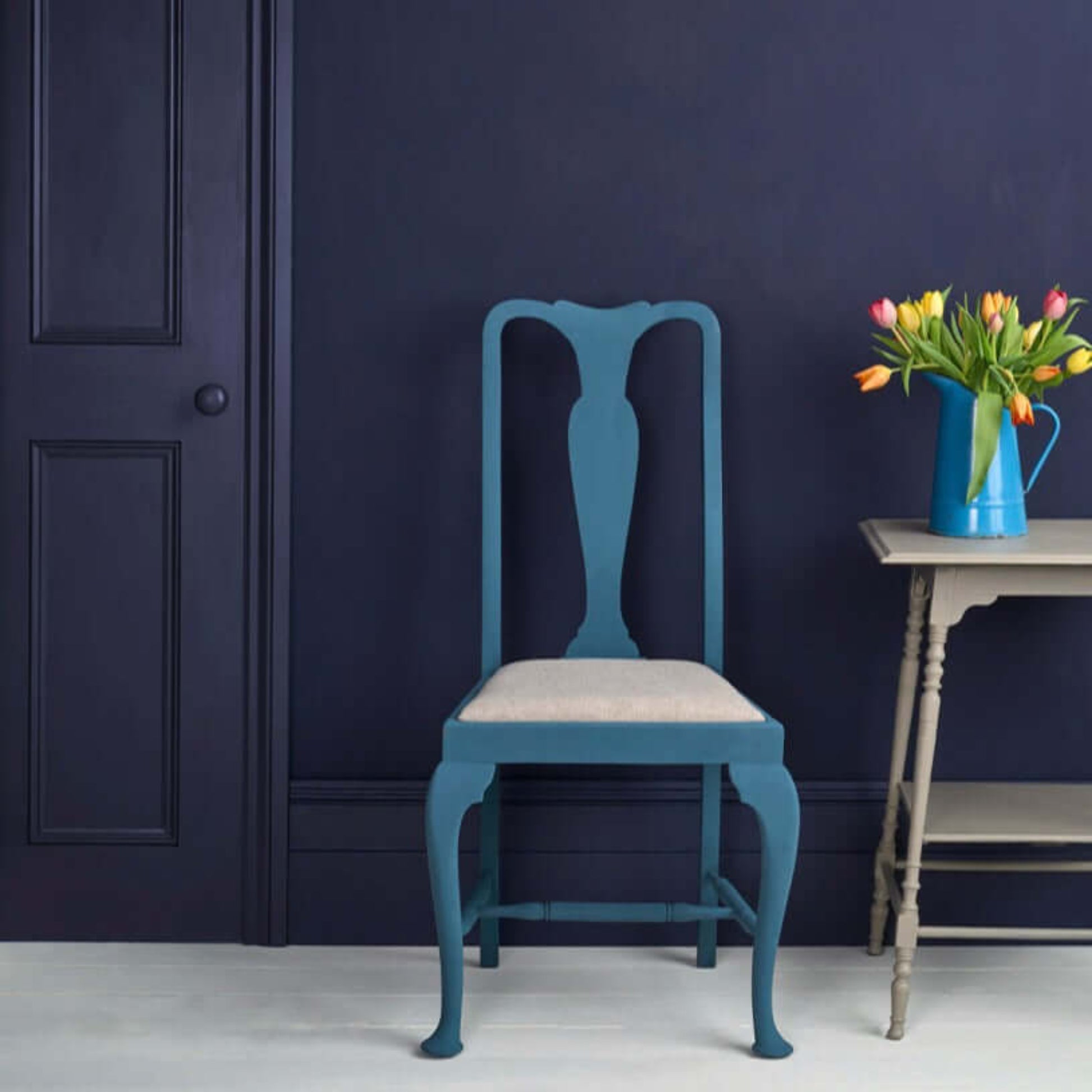 A stylish chair painted in Oxford Navy - Annie Sloan Satin Paint against a dark wall, with a vase of tulips.