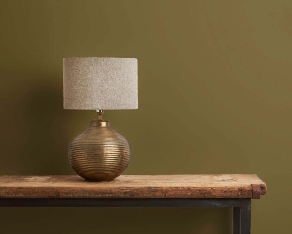 Bronze table lamp with a beige shade on a rustic wooden table against an olive green wall.