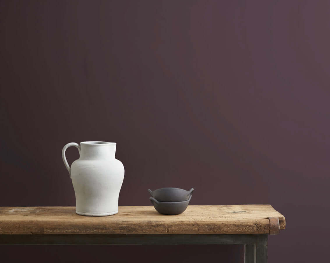 Tyrian Plum - Annie Sloan Wall Paint® used as a backdrop for a white pitcher and dark bowl on a wooden table.