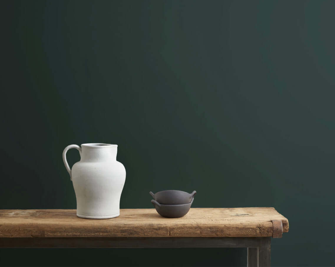 Knightsbridge Green - Annie Sloan Wall Paint showcases a white pitcher and a dark bowl on a wooden table against a green wall.