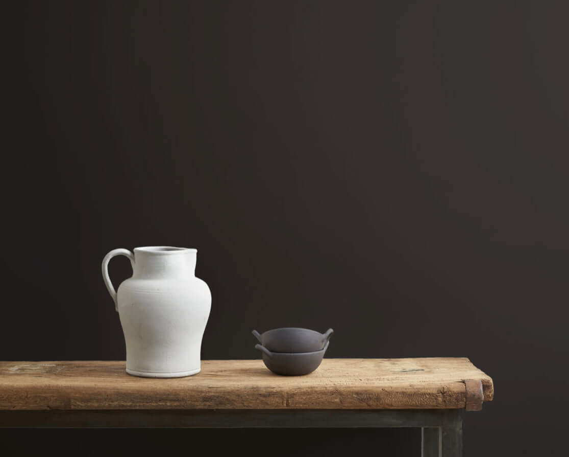 A white ceramic pitcher and a small dark bowl on a rustic wooden table against a dark wall.